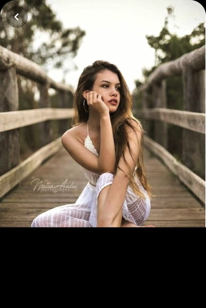 A young woman with long brown hair sits on a wooden bridge, wearing a white dress and looking thoughtfully to the side.