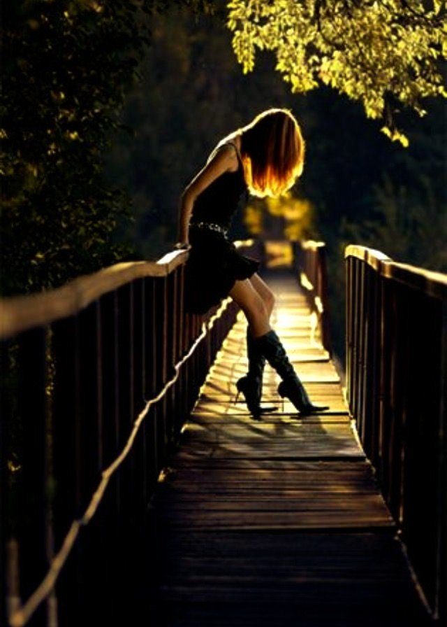 A woman in a black dress and high boots sits on a wooden bridge at night, illuminated by warm light.