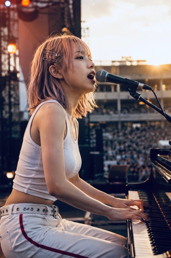 A young woman with pink hair, wearing a white tank top and white pants, plays the piano and sings into a microphone on a stage during golden hour.