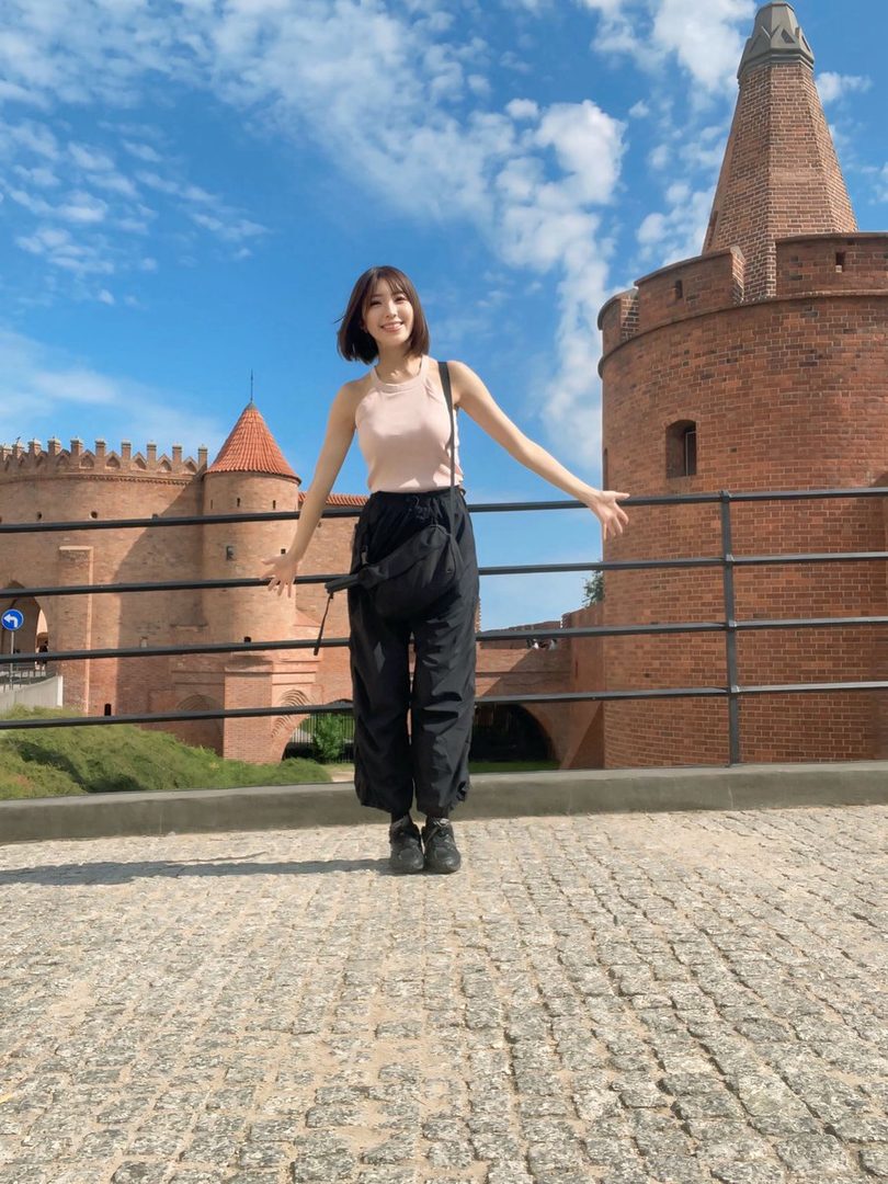 A young woman with short black hair smiles at the camera, standing outdoors in front of a historic brick building with towers under a blue sky with clouds.