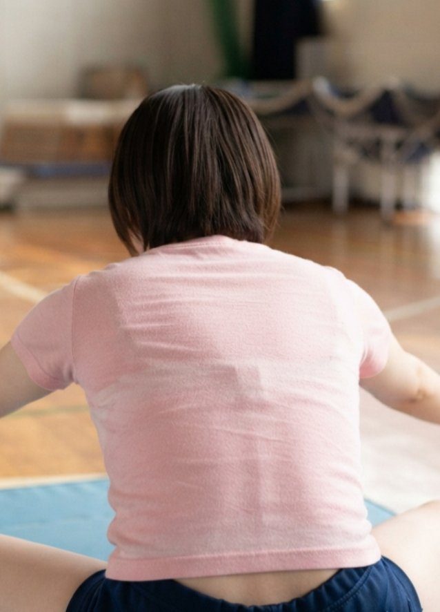 A person with short dark hair seen from behind, wearing a pink t-shirt and dark shorts, sitting on a blue mat in an indoor setting.