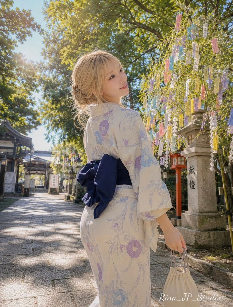 A young Japanese woman with blonde hair tied up, wearing a light-colored floral yukata and a dark blue obi, poses outdoors in a temple or shrine setting with trees and colorful Tanabata decorations.