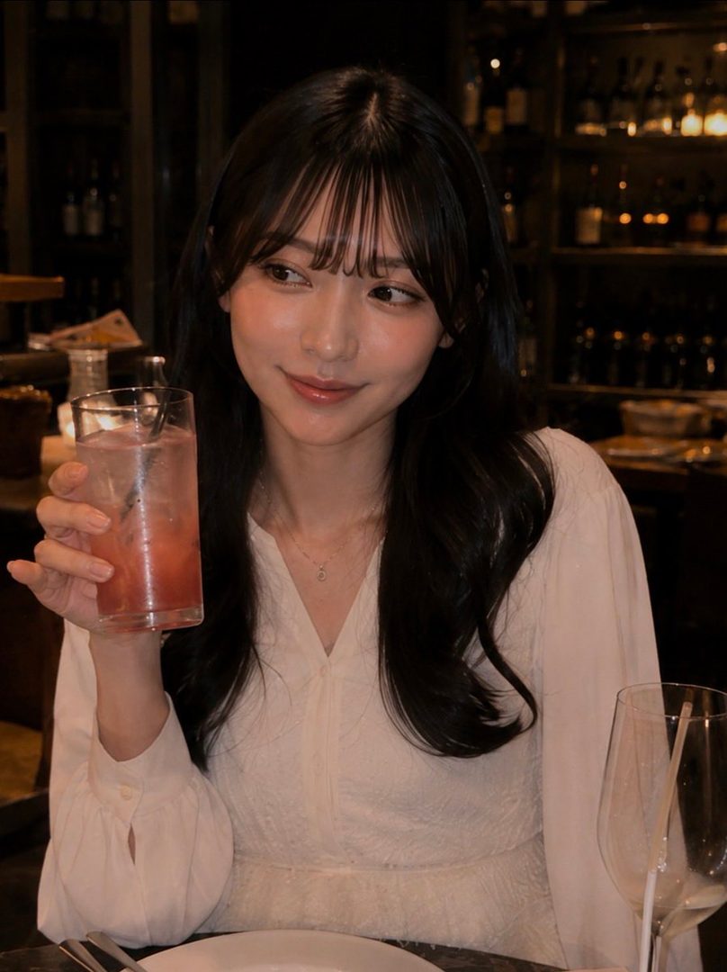 A young East Asian woman with long black hair and bangs, wearing a white dress, holds a pink drink and smiles gently in a dimly lit indoor setting.