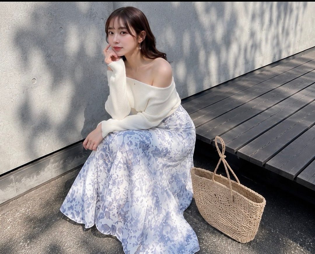 A young East Asian woman with long brown hair, wearing an off-the-shoulder white sweater and a floral print skirt, poses outdoors with a straw bag.