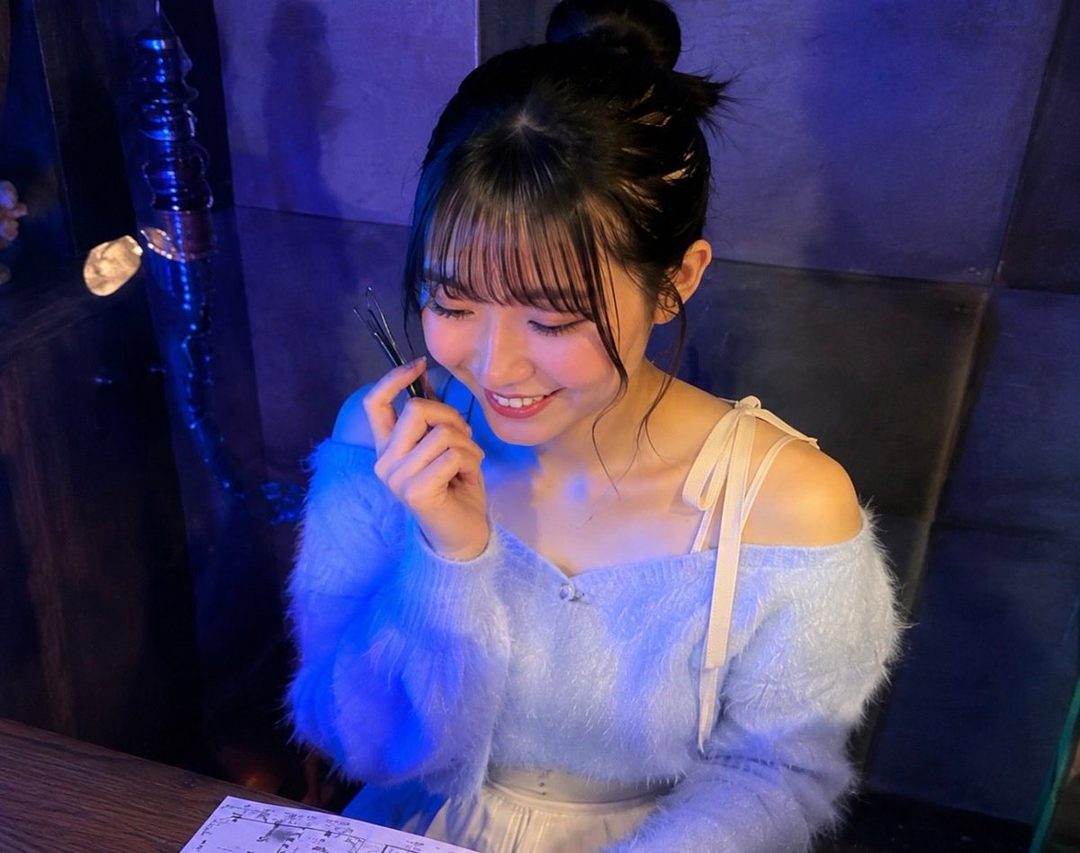 A young East Asian woman with her hair in a bun, wearing a light blue fuzzy off-the-shoulder sweater, smiles while holding a pen.