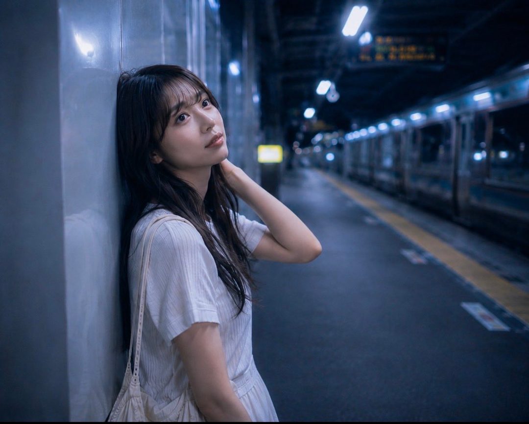 A young Japanese woman with long dark hair leans against a wall at a train station platform at night, looking thoughtful.