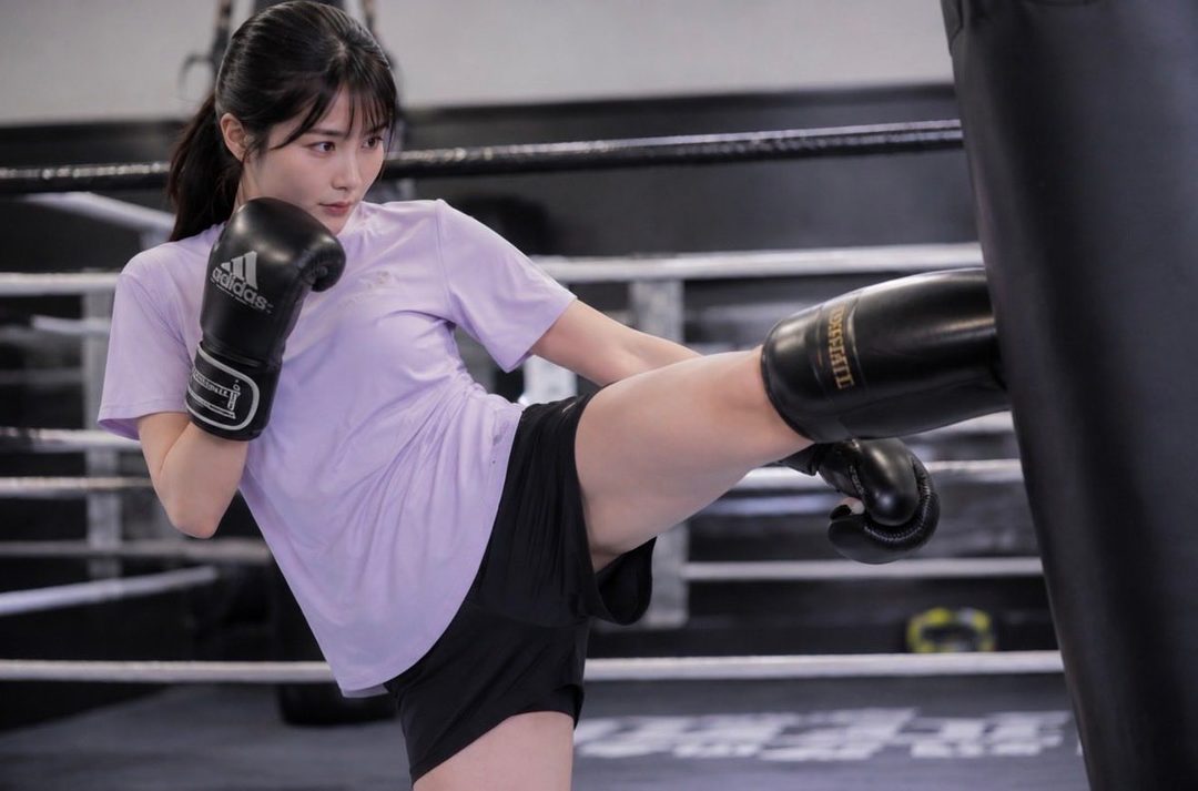 A young East Asian woman with black hair in a ponytail, wearing a purple t-shirt and black shorts, kicks a punching bag in a gym. She is wearing boxing gloves.