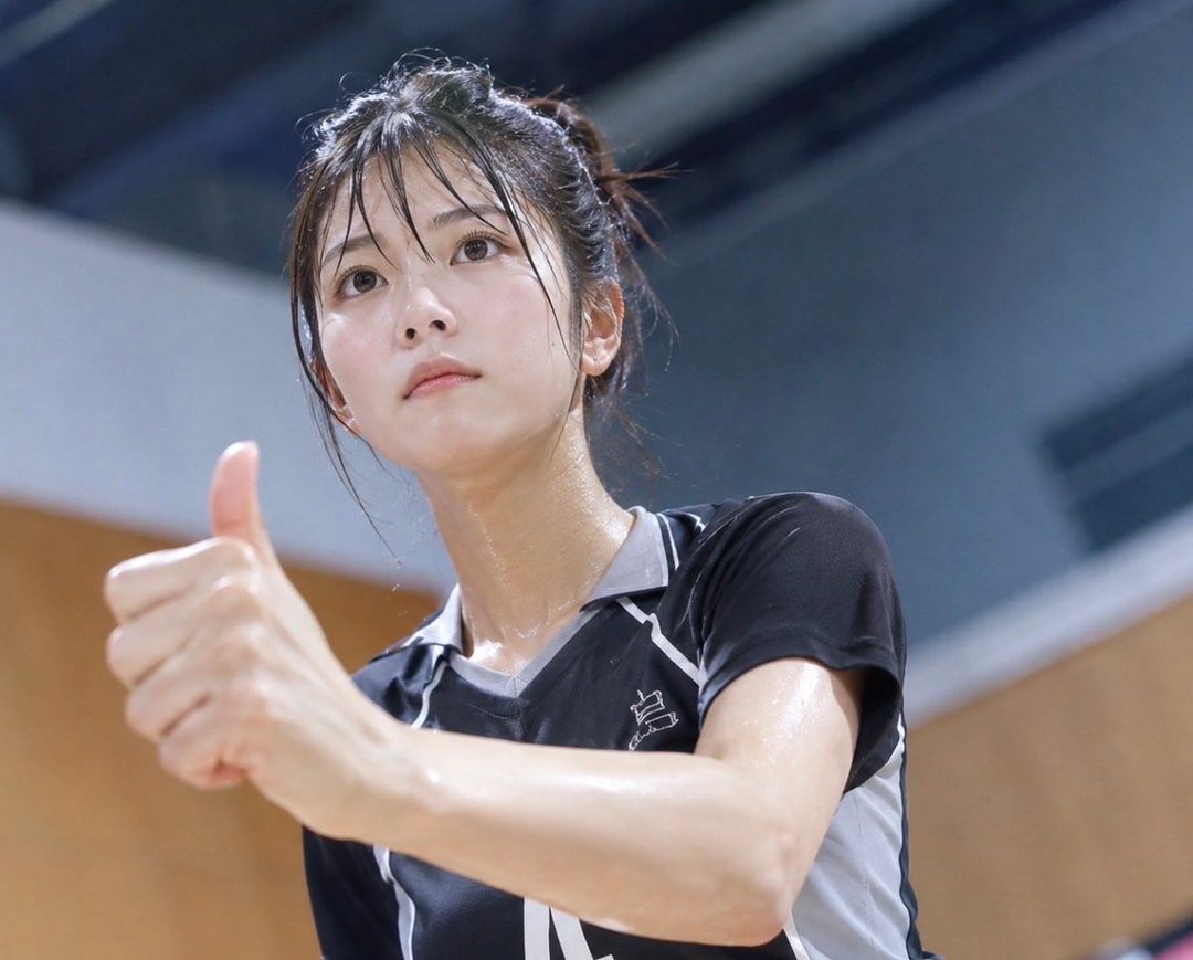 A young East Asian woman with her hair in a ponytail, wearing a black and grey sports jersey, gives a thumbs up with a sweaty face, indoors.