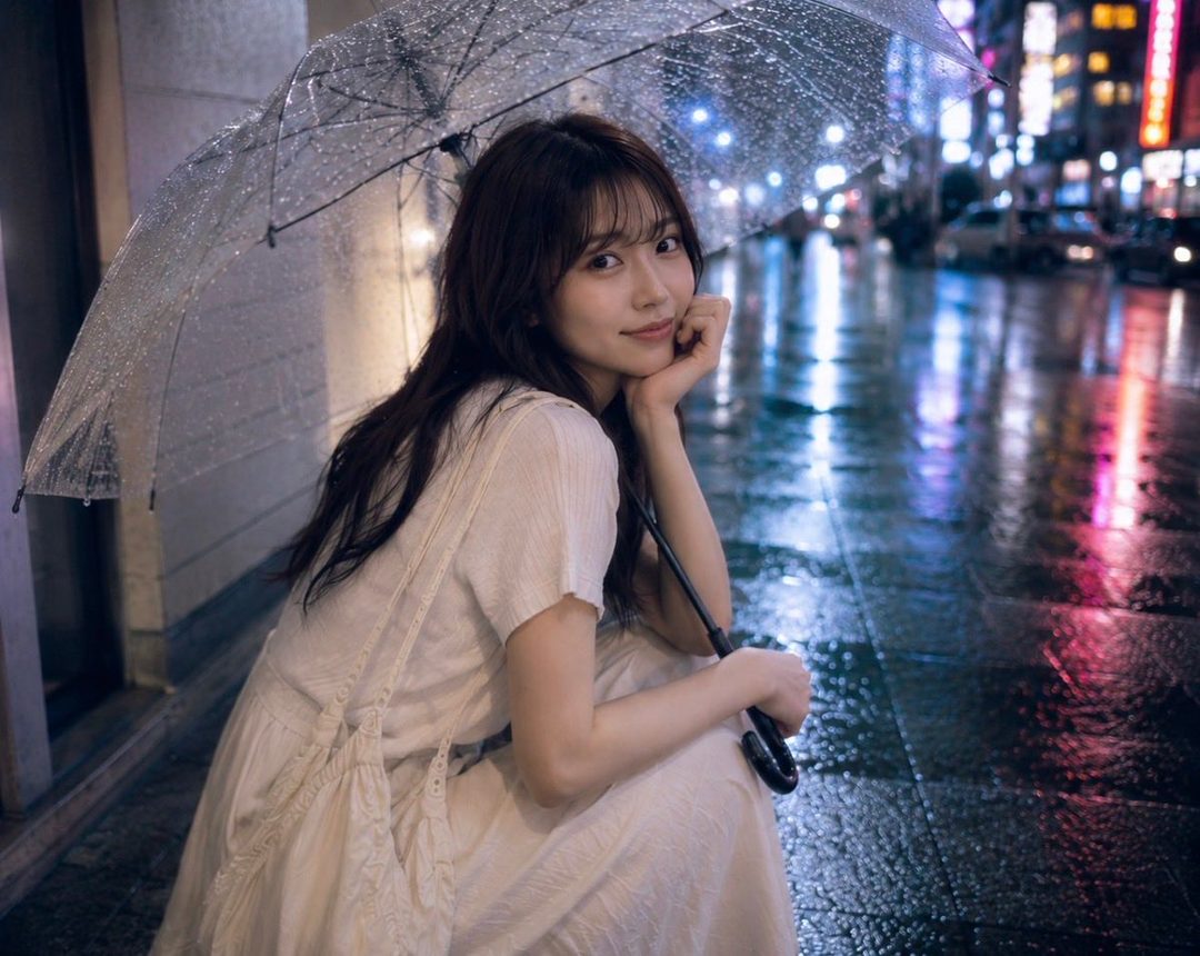 A young woman with long brown hair sits under a clear umbrella on a rainy city street at night, looking at the camera with a gentle smile.