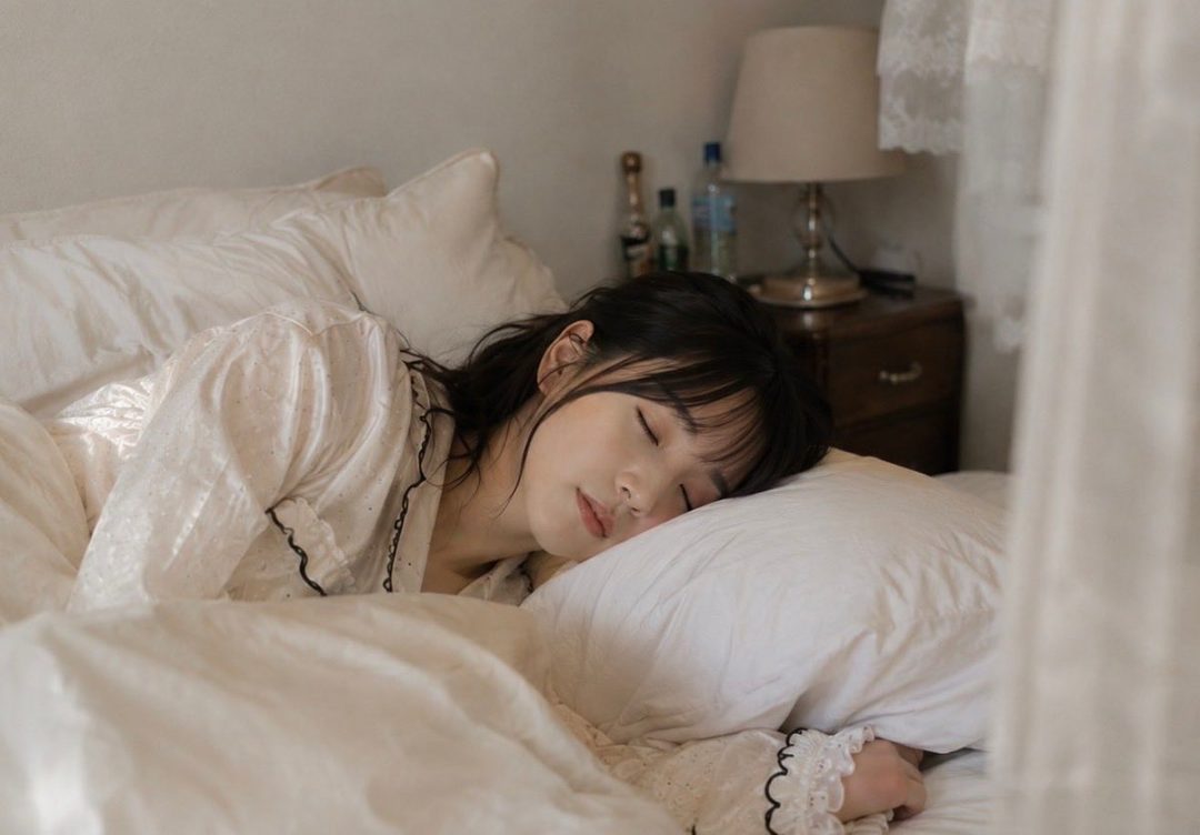 A young woman with black hair sleeps peacefully in a bed, wearing a white pajama top.