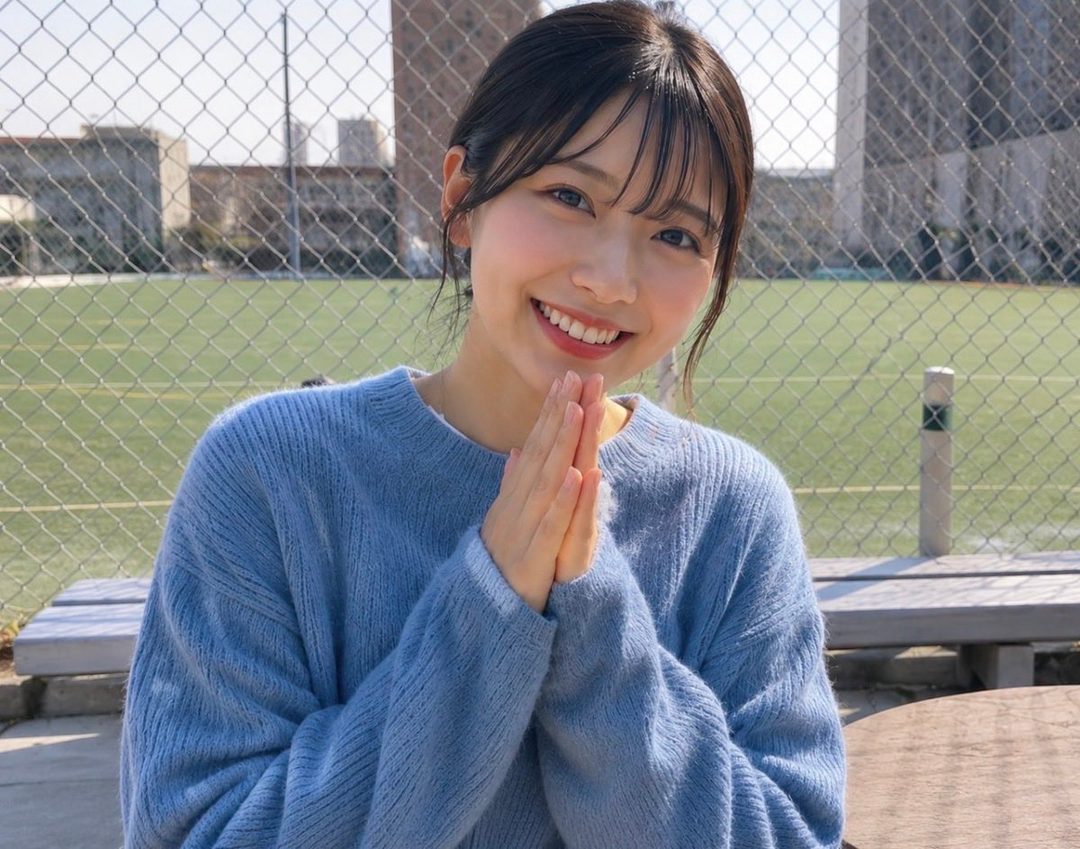 A young East Asian woman with dark hair and bangs, wearing a light blue sweater, smiles warmly with her hands clasped in front of her face outdoors.