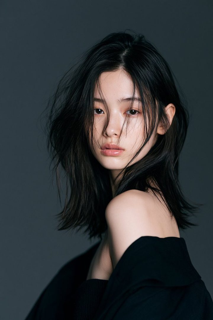 A studio portrait of a young East Asian woman with long, dark, windswept hair, wearing a black off-the-shoulder top against a dark grey background.
