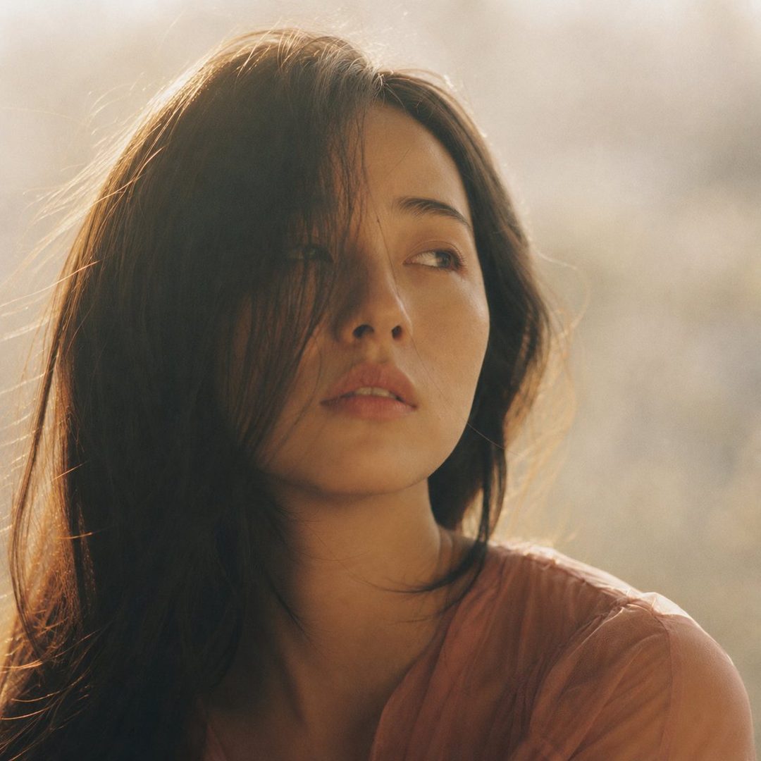 Close-up portrait of a young Japanese woman with dark hair partially obscuring her face, looking dreamily into the distance, wearing a dusty rose blouse, with soft, hazy afternoon light.
