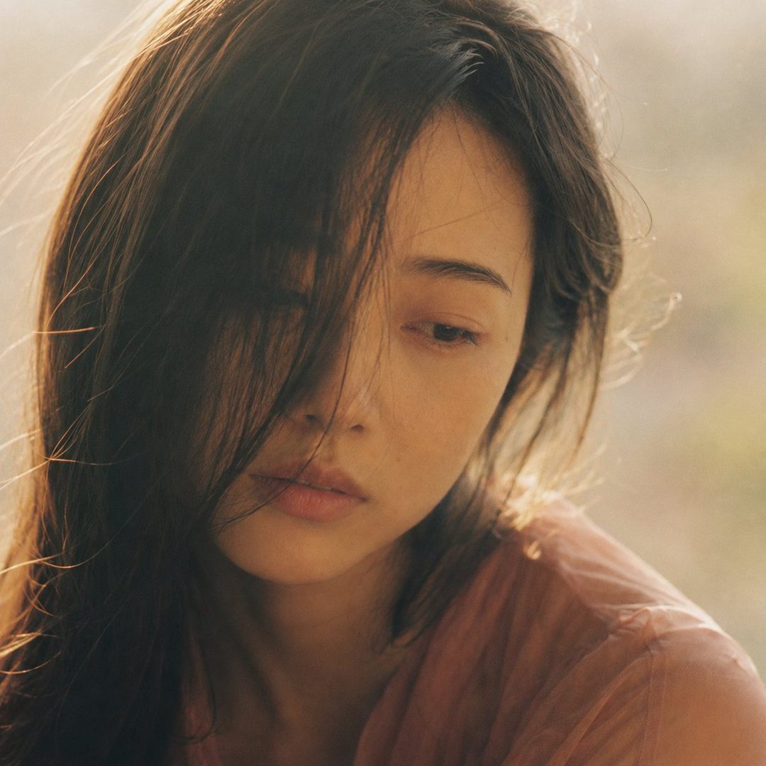 Close-up portrait of a young Japanese woman with dark hair partially obscuring her face, looking dreamily into the distance with soft, hazy afternoon light.