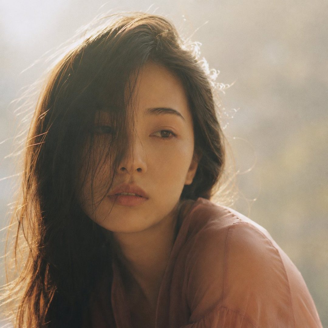 Close-up portrait of a young Japanese woman with long dark hair partially obscuring her face, wearing a dusty rose blouse, with a dreamy expression and soft, hazy lighting.
