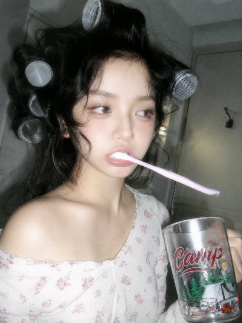 A young woman with hair curlers in her hair holds a toothbrush in her mouth and a cup in her hand, looking off to the side.