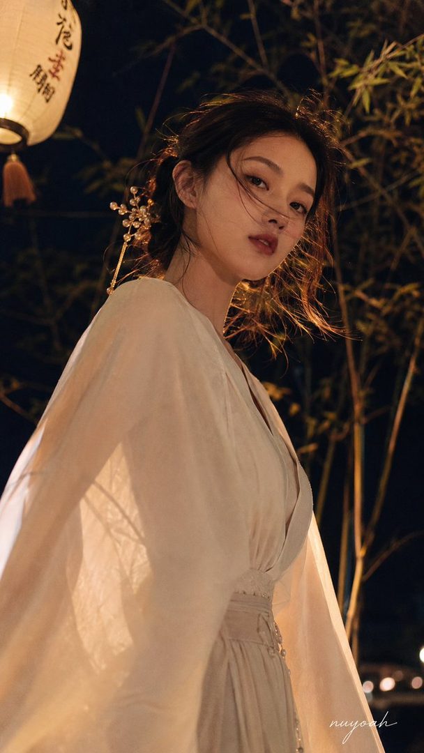 A young East Asian woman with dark hair styled with an ornate hairpin, wearing a flowing light-colored dress, poses outdoors at night with a traditional lantern and bamboo in the background.