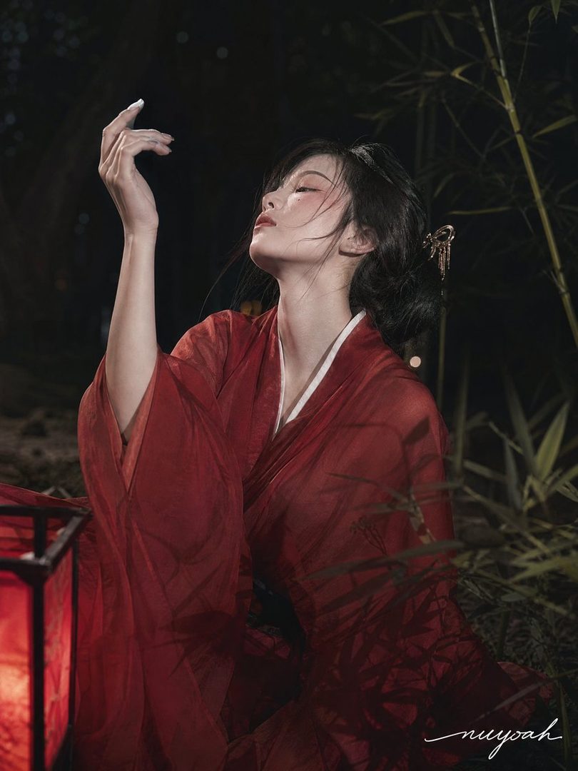 A woman in a red traditional Chinese dress poses gracefully in a dimly lit outdoor setting with bamboo and a lantern.