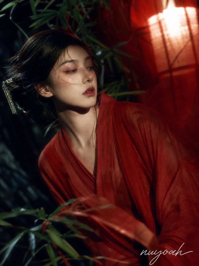 A young East Asian woman with dark hair, wearing a flowing red robe, poses elegantly outdoors at night, with soft lantern light illuminating her face and the fabric.
