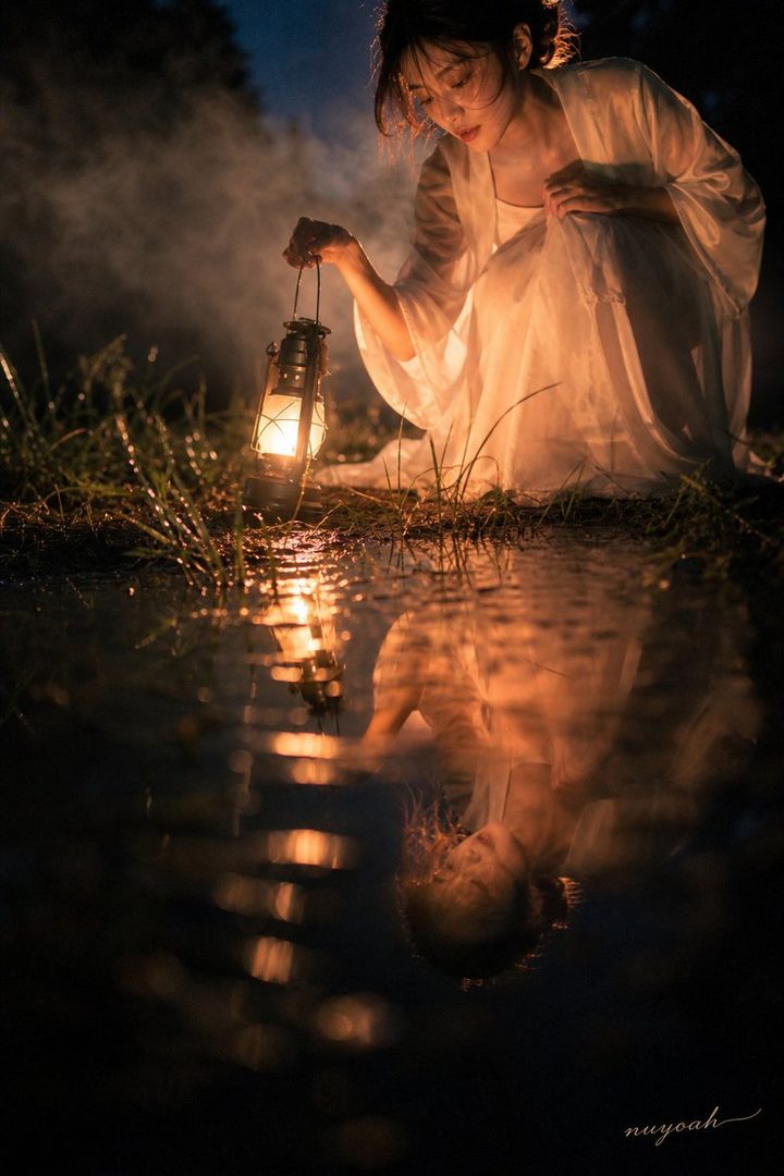 A young woman in a white dress holds a lantern, her reflection visible in the water.