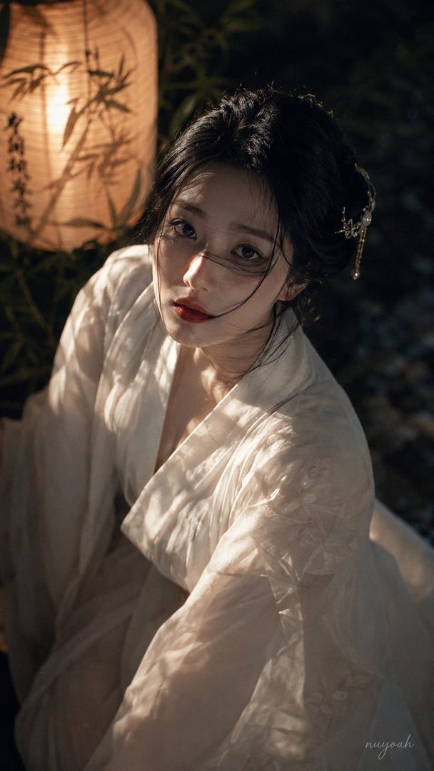 A beautiful East Asian woman with black hair in a traditional white robe, illuminated by a warm lantern light, with bamboo shadows in the background.