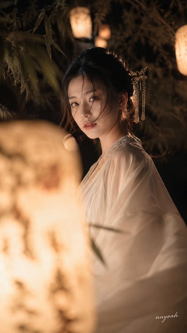 A beautiful East Asian woman with dark hair styled up, wearing a flowing white robe, poses in a dimly lit setting with lanterns and bamboo shadows, exuding a dreamy and elegant mood.
