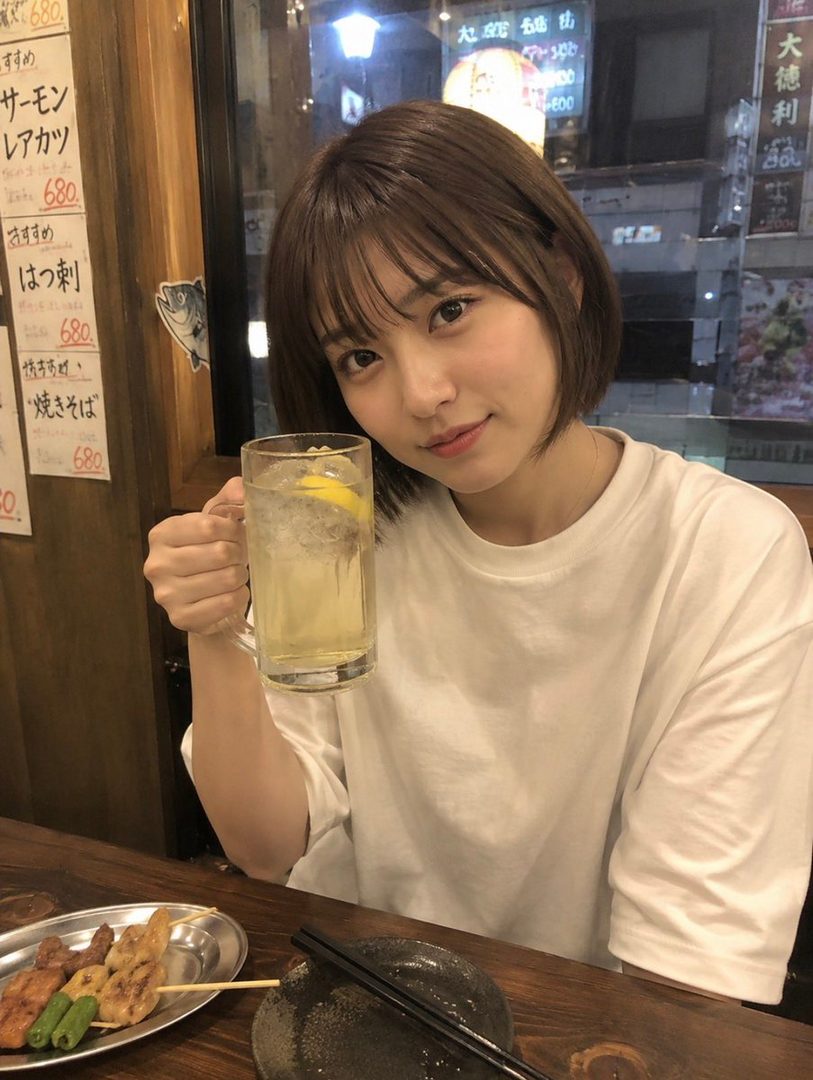 A young woman with short brown hair and bangs, wearing a white t-shirt, holds a glass of a lemon drink and smiles at the camera. She is sitting at a table in a restaurant with skewers of food in front of her.