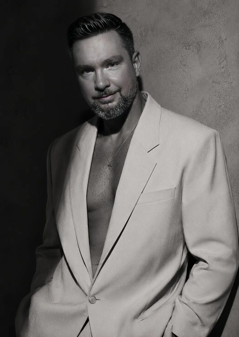 A man in an open suit jacket with a necklace, posing against a textured wall.
