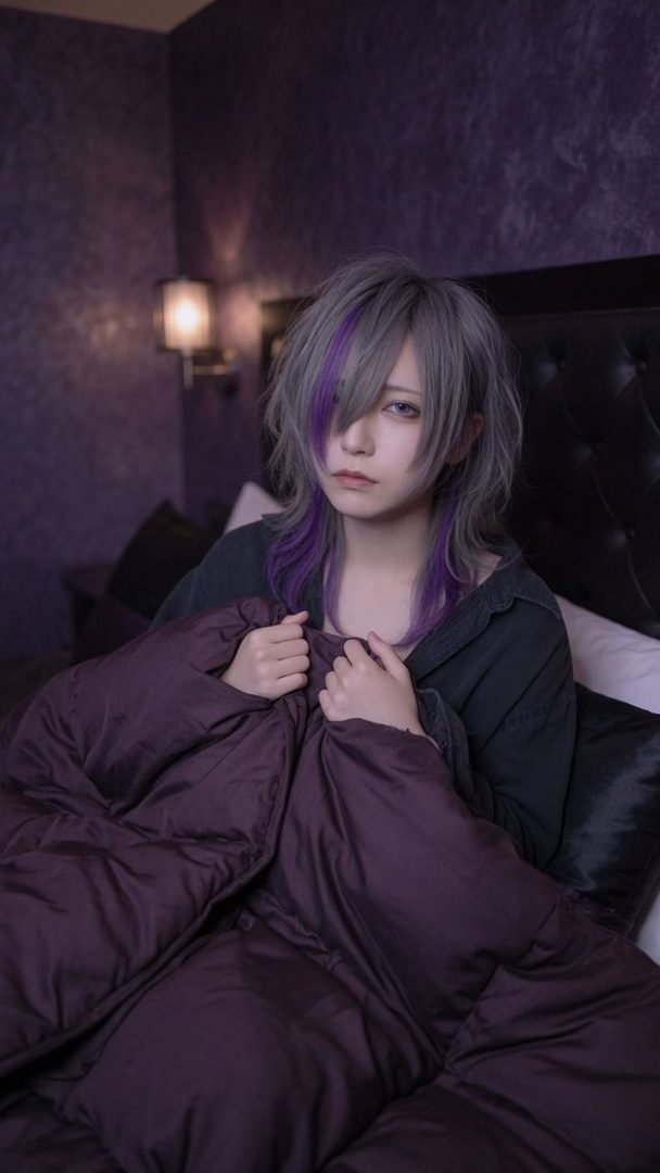 A person with long, gray hair with purple streaks, wrapped in a dark purple blanket, looking at the camera with a gentle expression.