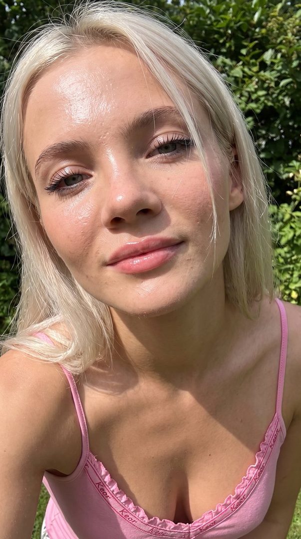 Close-up selfie of a young woman with blonde hair and a pink top, outdoors with greenery in the background.
