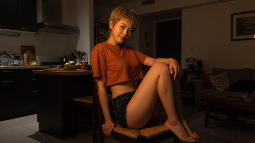 A young East Asian woman with short blonde hair, wearing a cropped orange t-shirt and denim shorts, sits on a chair in a dimly lit room, posing playfully.