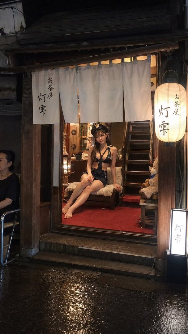 A photorealistic AI-generated portrait of a young woman in a police-themed outfit, sitting indoors in front of a traditional Japanese establishment at night.