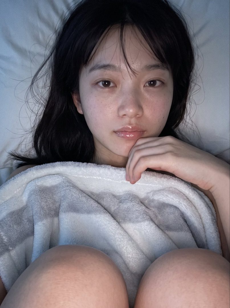A young East Asian woman with black hair and freckles, lying in bed and looking at the camera with a hand on her chin, wrapped in a blanket.