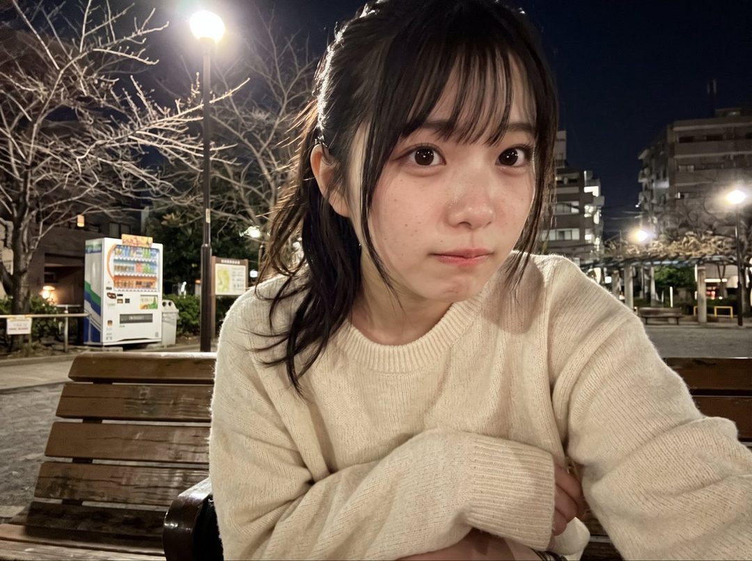 A young Japanese woman with dark hair and bangs, wearing a cream-colored sweater, takes a selfie at night in a park with a vending machine and streetlights visible.