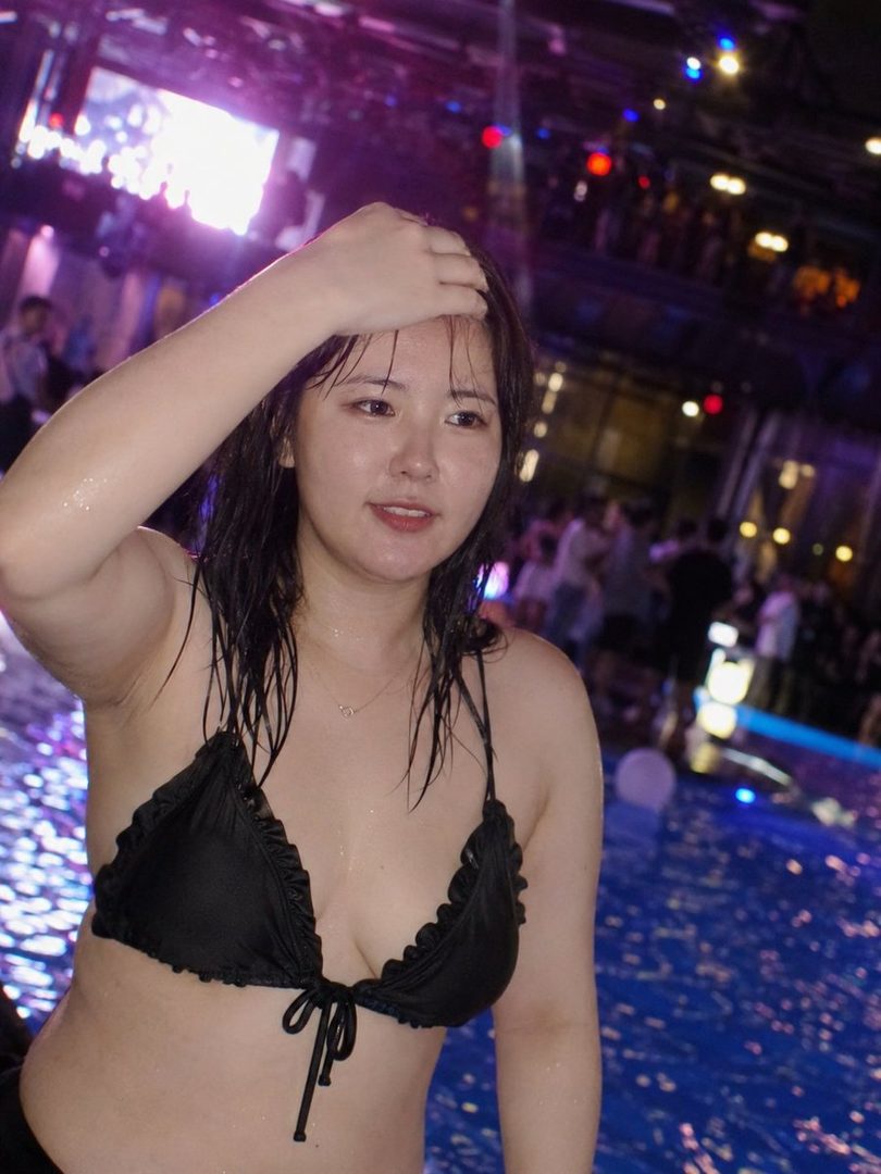 A young woman in a black bikini poses by a pool at night, with colorful lights in the background.