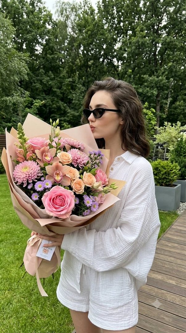 A young woman in a white linen outfit and sunglasses holds a large bouquet of pink and peach flowers, smelling them in a garden setting.