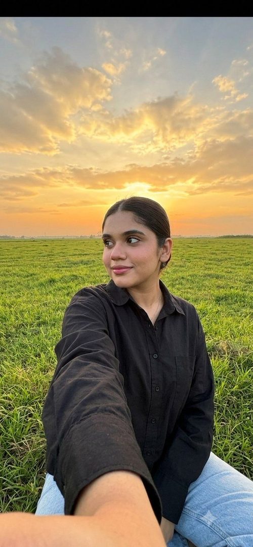 A young woman takes a selfie in a green field during golden hour, looking to the side with a gentle smile.