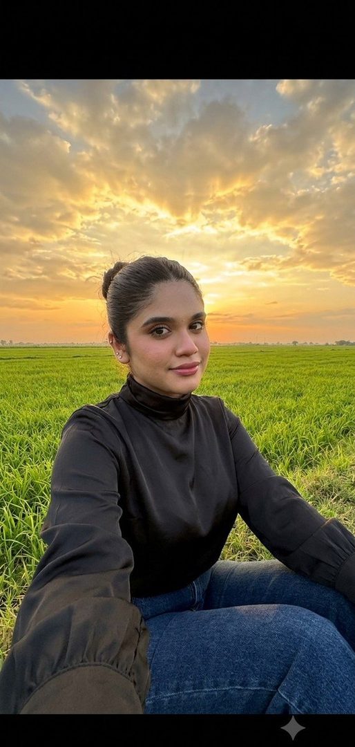 A young woman with dark hair in a bun takes a selfie in a green field at sunset, wearing a black top and blue jeans.