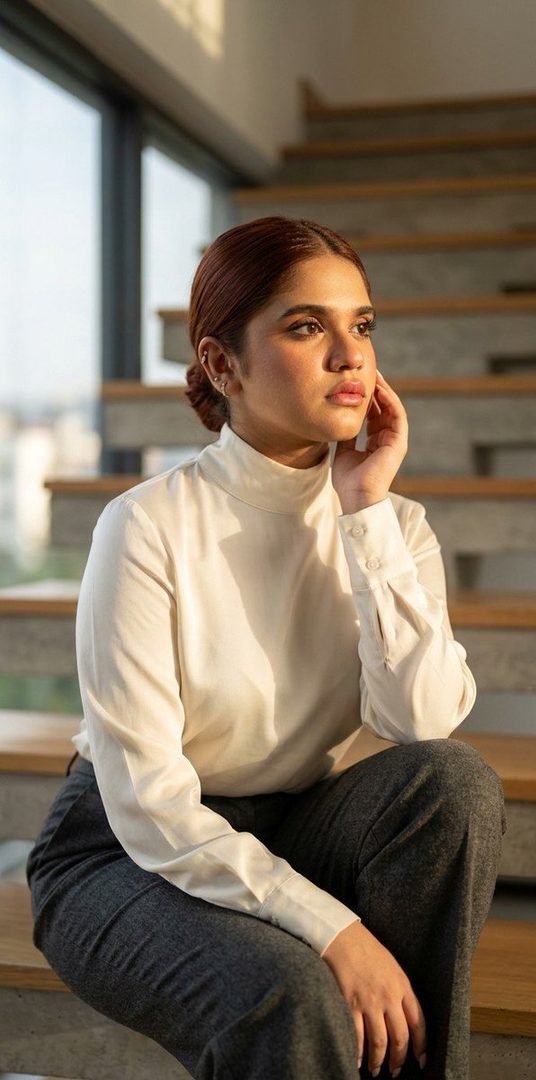 A woman with brown hair in a white turtleneck blouse and dark pants sits on a modern staircase, looking thoughtfully into the distance during golden hour.