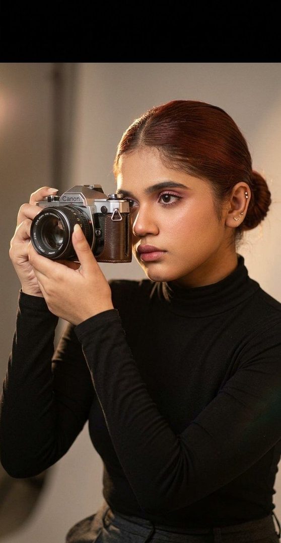 A young woman with brown hair in a black turtleneck holds a vintage Canon camera to her eye in a studio setting.