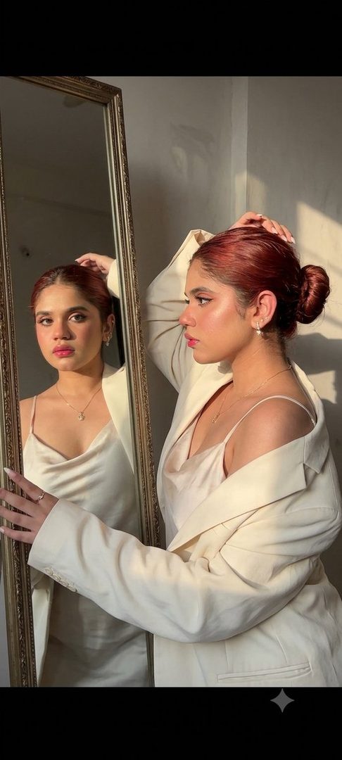 A woman with red hair in a bun, wearing a white camisole and blazer, looks into a mirror with sunlight casting shadows on the wall.