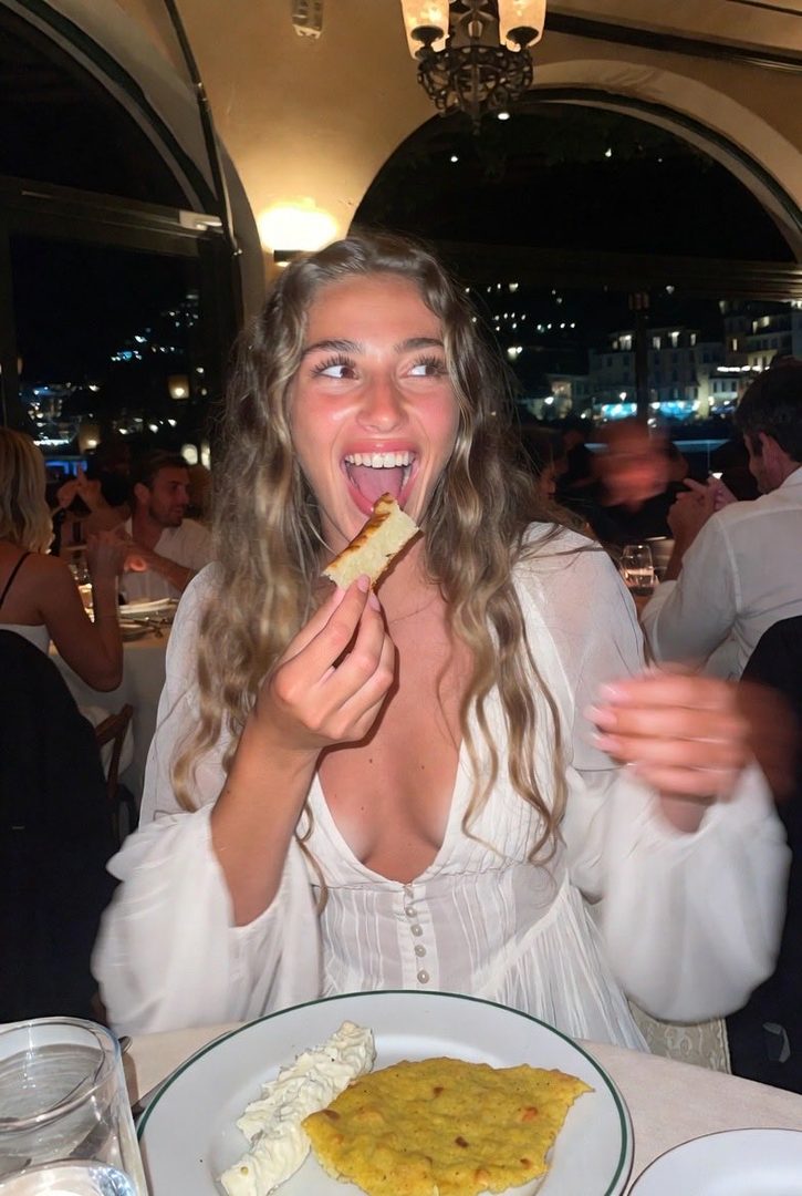 A young woman with long blonde hair, wearing a white dress, is joyfully eating a piece of food at a restaurant table at night.