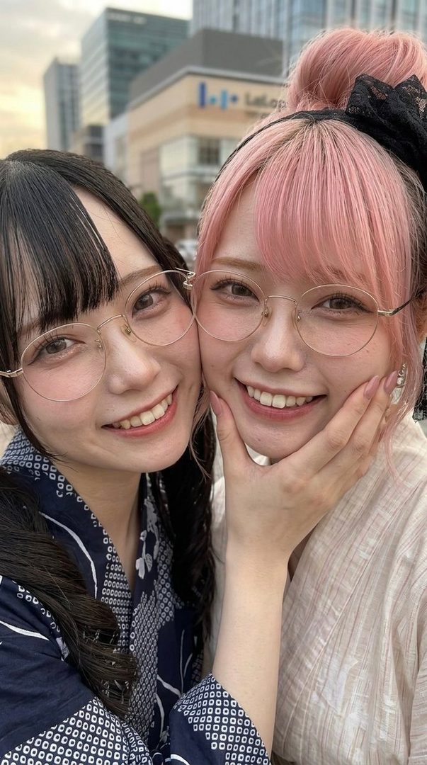 Two young Japanese women with glasses and yukatas smiling at the camera, one with pink hair and the other with black hair.