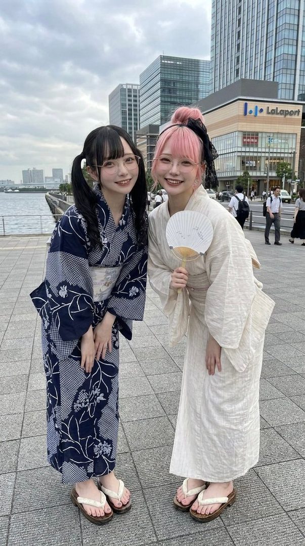 Two young women in yukatas, one with black pigtails and glasses, the other with pink hair and a fan, posing outdoors in a city with modern buildings and a waterfront.