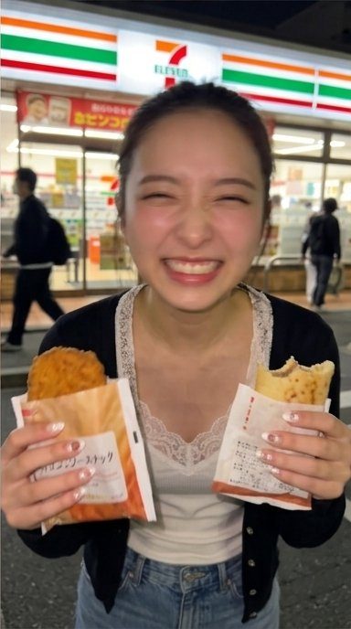A young East Asian woman with a bright smile and expressive eyes, holding two convenience store snacks in front of a 7-Eleven store at night.