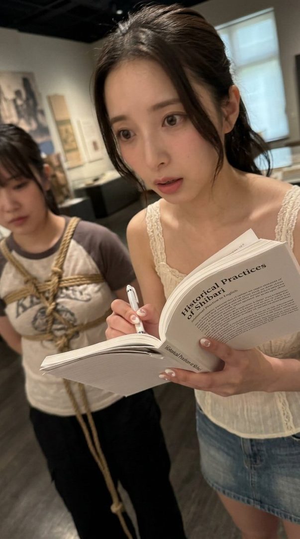 A young woman with long dark hair, wearing a white top and denim skirt, reads a book titled 'Historical Practices of Shibari'. Another woman with dark hair is visible behind her, wearing a t-shirt and black pants, with rope binding her torso.