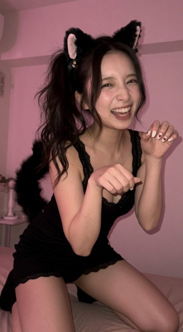 A young woman with cat ears and a tail smiles playfully, wearing a black lace-trimmed dress indoors.