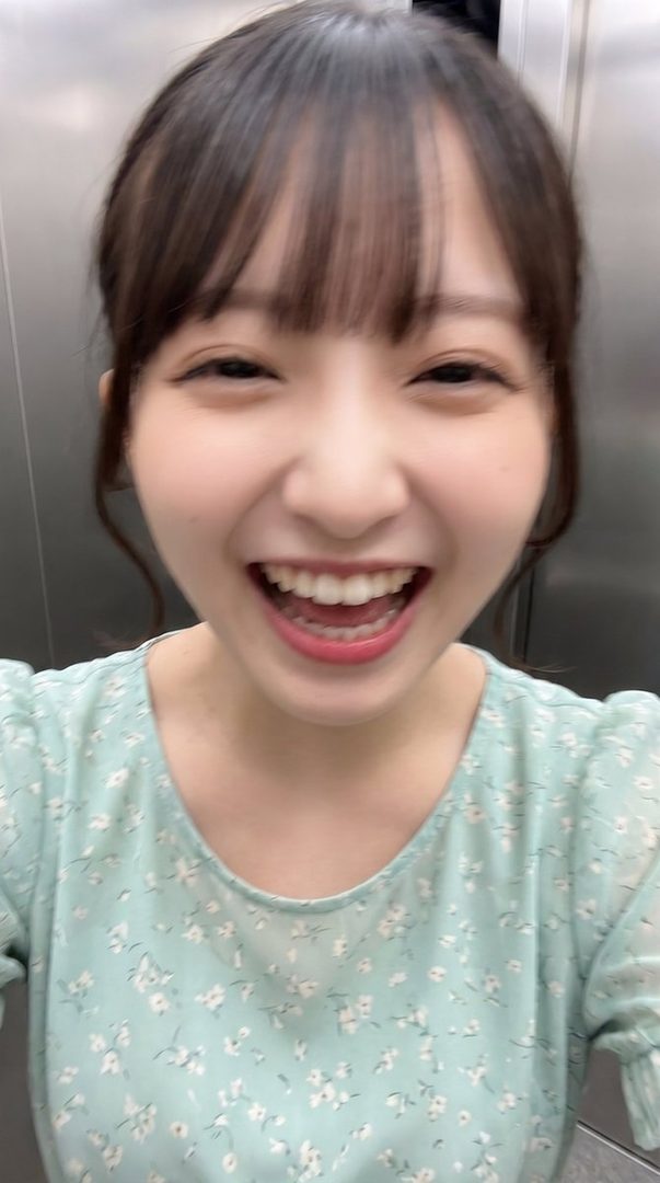 A young woman with brown hair and bangs smiles widely in a selfie taken indoors, wearing a light blue floral print top.