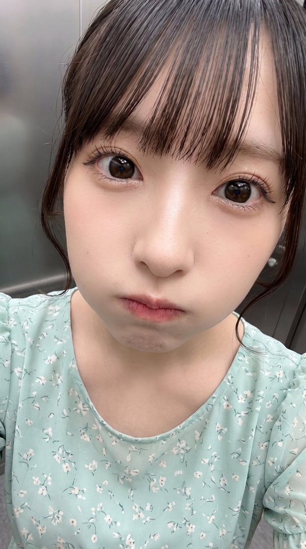 A young East Asian woman with dark hair and bangs, wearing a light blue floral blouse, takes a selfie with a pouty expression.