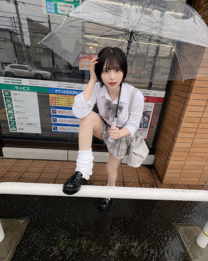 A young Japanese woman in a school uniform poses under a clear umbrella on a rainy day, holding her hand to her head.